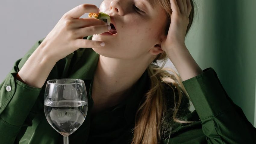 Portrait of a young woman savoring sushi with a wine glass in a relaxed, green-themed setting.